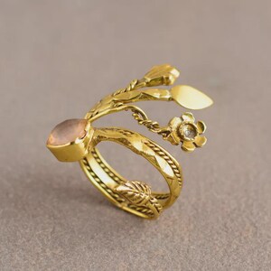 Peut inclure: Une bague dor&eacute;e orn&eacute;e d'une pierre rose clair et d'un motif floral. La bague a un anneau d&eacute;taill&eacute; et textur&eacute; avec des fleurs et des feuilles. La pierre pr&eacute;cieuse est sertie dans un sertissage carr&eacute;.