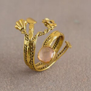 Peut inclure: Une bague dor&eacute;e avec un motif floral et de feuilles. La bague pr&eacute;sente une pierre pr&eacute;cieuse rose clair de coupe carr&eacute;e. La bande a un motif tress&eacute; textur&eacute;. La bague a un design ouvert unique.