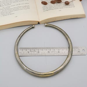 May include: A silver-toned choker necklace with a ribbed design. The necklace is approximately 30 cm in diameter. The ends of the necklace have small, rounded silver accents. A ruler is placed near the necklace.