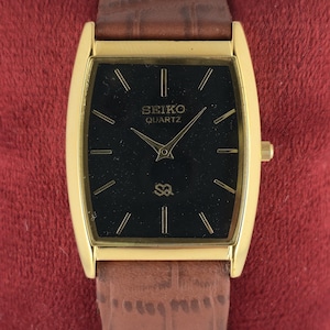 Puede incluir: Un reloj Seiko Quartz dorado con una esfera negra rectangular y agujas doradas. El reloj tiene una correa de cuero marrón. La esfera del reloj muestra el logotipo de Seiko Quartz y marcadores de hora.