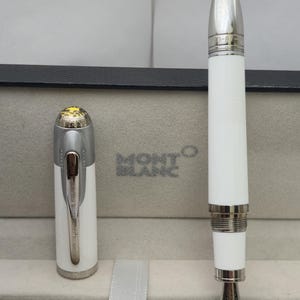 Könnte beinhalten: Ein weißer Montblanc Füllfederhalter mit silbernen Akzenten, präsentiert in seinem Etui. Der Stift hat eine silberne Feder und eine Kappe mit dekorativem Design. Ein luxuriöses Schreibgerät.