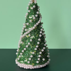 May include: A beaded Christmas tree decoration. The tree is cone-shaped and covered in green beads, with white pearl-like beads scattered throughout. A strand of white beads wraps around the tree. The base and top are trimmed with white beads.