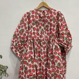 May include: A white dress with a red tomato pattern, hanging on a wooden hanger. The dress features long sleeves, a tie at the neckline, and a tiered skirt. The tomatoes have green leaves.