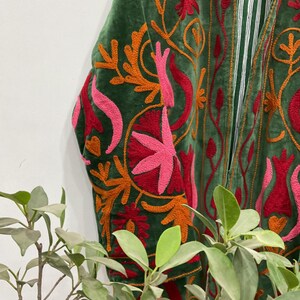 以下が含まれることがあります： オレンジ、赤、ピンクの色合いで、精巧な花の刺繍が施された緑色のベルベットジャケットのクローズアップ。 ジャケットの内側はストライプ模様です。 袖と前面が見え、詳細な職人技が示されています。