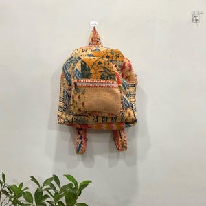 Puede incluir: Una mochila estampada y colorida con un bolsillo frontal y correas ajustables. La mochila presenta una mezcla de tela naranja, azul y amarilla con una textura acolchada. Cuelga de una pared blanca.