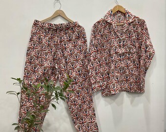 May include: A two-piece pyjama set with a floral pattern in brown, red, and white. The set includes a button-down shirt with a chest pocket and drawstring trousers. The pyjamas are hanging on wooden hangers against a white wall.