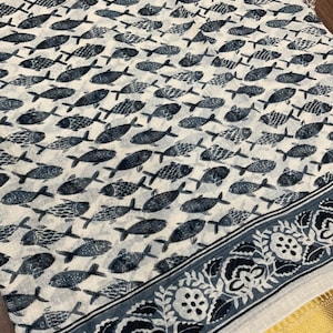 May include: Off-white fabric with a repeating pattern of stylized blue-gray fish. A border with floral and geometric designs in blue-gray and gold accents the edge of the fabric. The fabric appears to be lightweight.