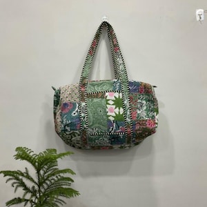 May include: A patchwork tote bag with a variety of floral and paisley patterns in shades of green, pink, and blue. The bag has a rectangular shape with a rounded top and two handles. The handles are green with a white and black stitched border.
