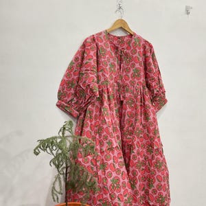 May include: A loose-fitting midi dress in a vibrant pink fabric featuring an all-over print of red tomatoes with green leaves. The dress has three-quarter length sleeves with gathered cuffs, a round neckline with a small tie closure, and a tiered skirt.