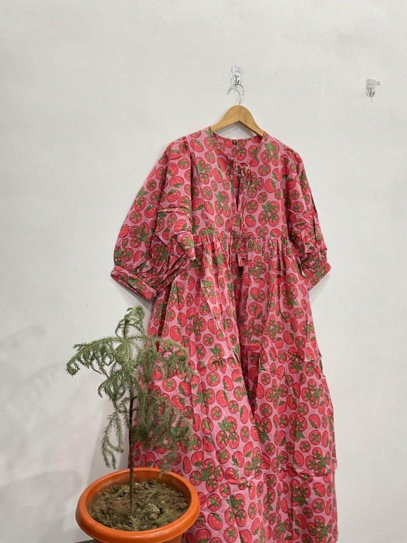 May include: A loose-fitting midi dress in a vibrant pink fabric featuring an all-over print of red tomatoes with green leaves. The dress has three-quarter length sleeves with gathered cuffs, a round neckline with a small tie closure, and a tiered skirt.