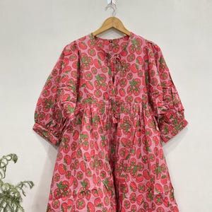 May include: A pink midi dress featuring an all-over print of red tomatoes with green stems and leaves. The dress has a round neckline with a front tie closure, three-quarter length puff sleeves with ruffled cuffs, and a gathered waistline.