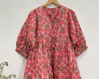 May include: A pink midi dress featuring an all-over print of red tomatoes with green stems and leaves. The dress has a round neckline with a front tie closure, three-quarter length puff sleeves with ruffled cuffs, and a gathered waistline.