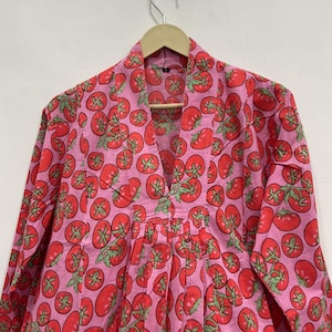 May include: A long-sleeved tunic featuring a vibrant pink background with a repeating pattern of red tomatoes and green stems. The garment has a V-neckline with a structured collar and vertical pleats extending from the center chest area.