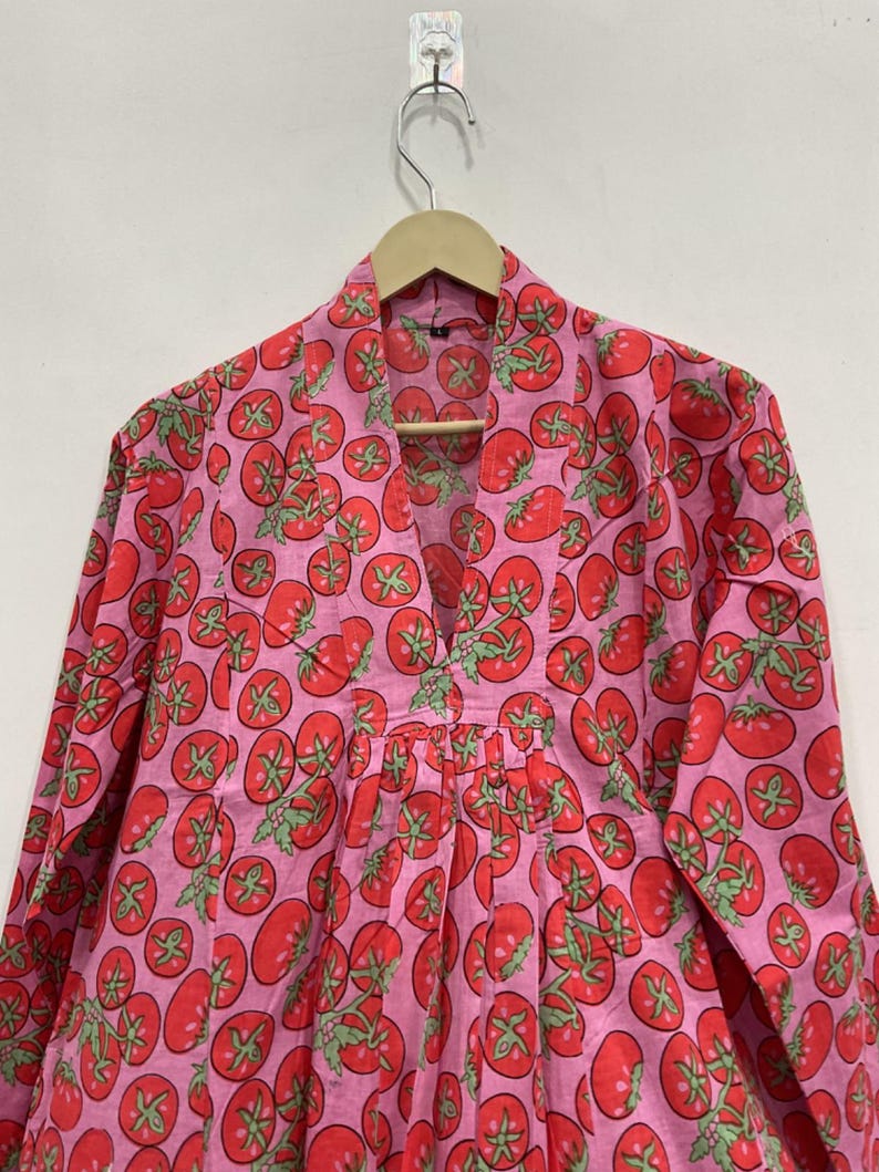 May include: A long-sleeved tunic featuring a vibrant pink background with a repeating pattern of red tomatoes and green stems. The garment has a V-neckline with a structured collar and vertical pleats extending from the center chest area.