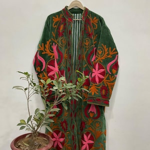 以下が含まれることがあります： ピンク、オレンジ、赤の色合いの花柄刺繍が施された、グリーンのベルベットのロングコート。木製のハンガーに掛けられています。コートの内側はストライプ柄です。鉢植えの植物が前景にあります。