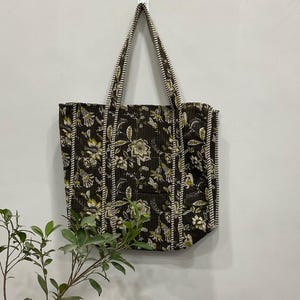 以下が含まれることがあります： 緑と白の色合いの花柄が施された、濃い茶色のトートバッグ。バッグには白い縦縞と長い持ち手が付いています。白い壁に掛けられています。