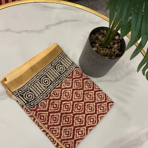 Könnte beinhalten: Ein gefaltetes Textil mit einem geometrischen Muster in Rot und Creme, mit einem schwarzen und cremefarbenen Rand. Das Textil liegt auf einer weißen Marmoroberfläche neben einer kleinen Topfpflanze.