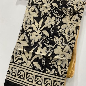 May include: A black and cream-colored scarf with a floral pattern and a decorative border. The scarf is draped over a wooden hanger. The floral design features large flowers and leaves. The border has a repeating geometric pattern.