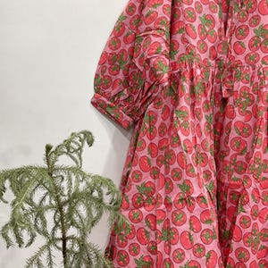 May include: A pink dress featuring a repeating pattern of red tomatoes with green stems and leaves. The dress has a round neckline with a tie closure, three-quarter length sleeves with cuffs, and a loose, flowing silhouette with gathered fabric.