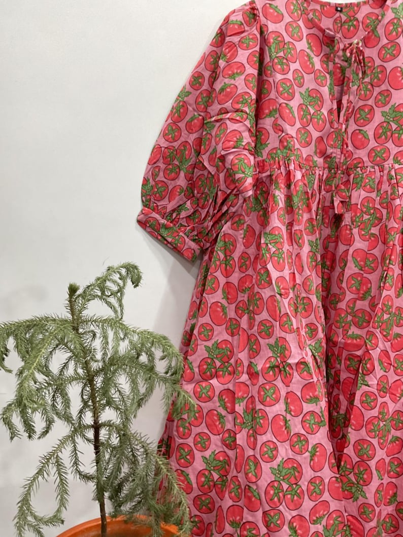 May include: A pink dress featuring a repeating pattern of red tomatoes with green stems and leaves. The dress has a round neckline with a tie closure, three-quarter length sleeves with cuffs, and a loose, flowing silhouette with gathered fabric.