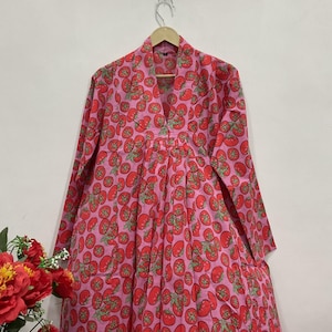 May include: A long-sleeved dress featuring a vibrant pink background with a repeating pattern of red tomatoes and green stems. The garment has a V-neckline, a structured collar, and a gathered waistline that flows into a full, pleated skirt.