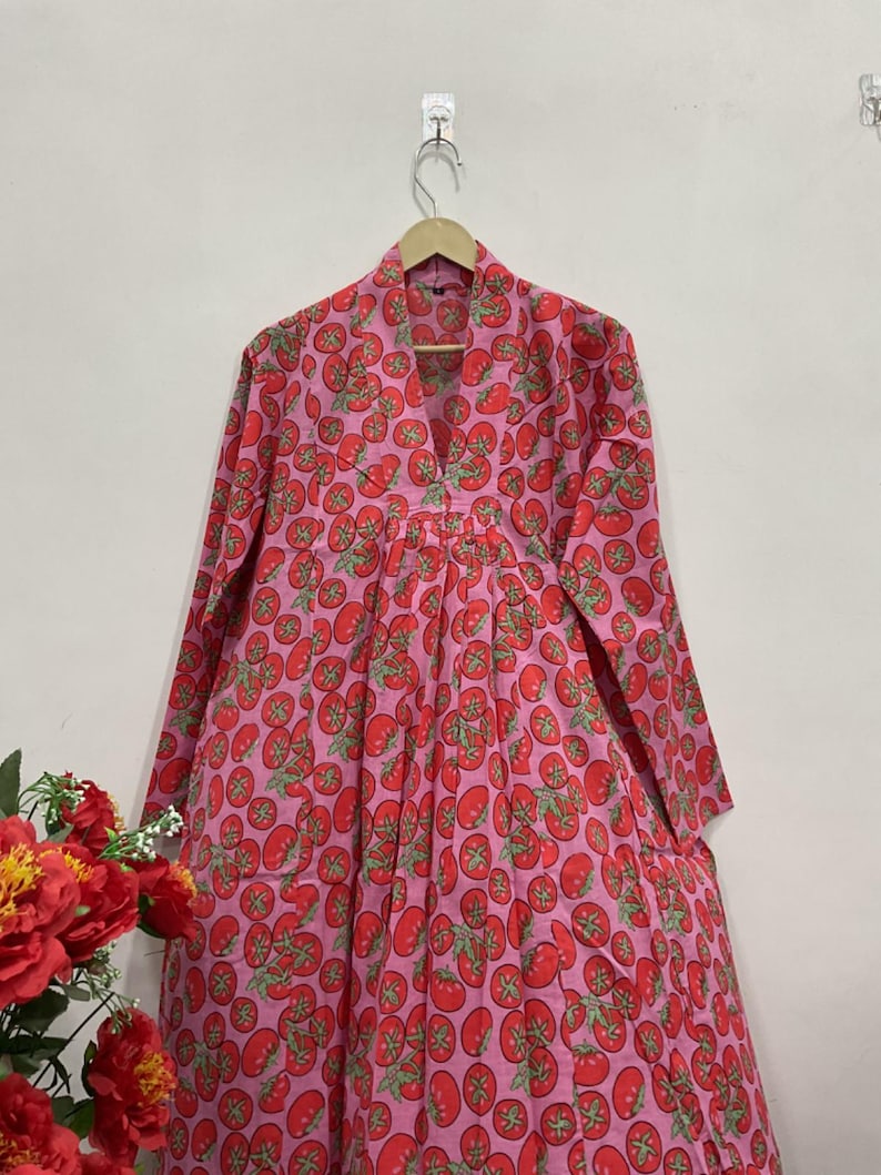 May include: A long-sleeved dress featuring a vibrant pink background with a repeating pattern of red tomatoes and green stems. The garment has a V-neckline, a structured collar, and a gathered waistline that flows into a full, pleated skirt.