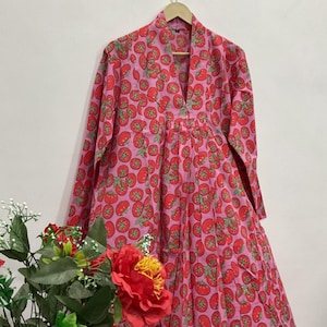 May include: A long-sleeved dress featuring a vibrant pink background with a repeating pattern of red tomatoes and green stems. The garment has a V-neckline, a structured collar, and a pleated bodice that flows into a full-length skirt silhouette.
