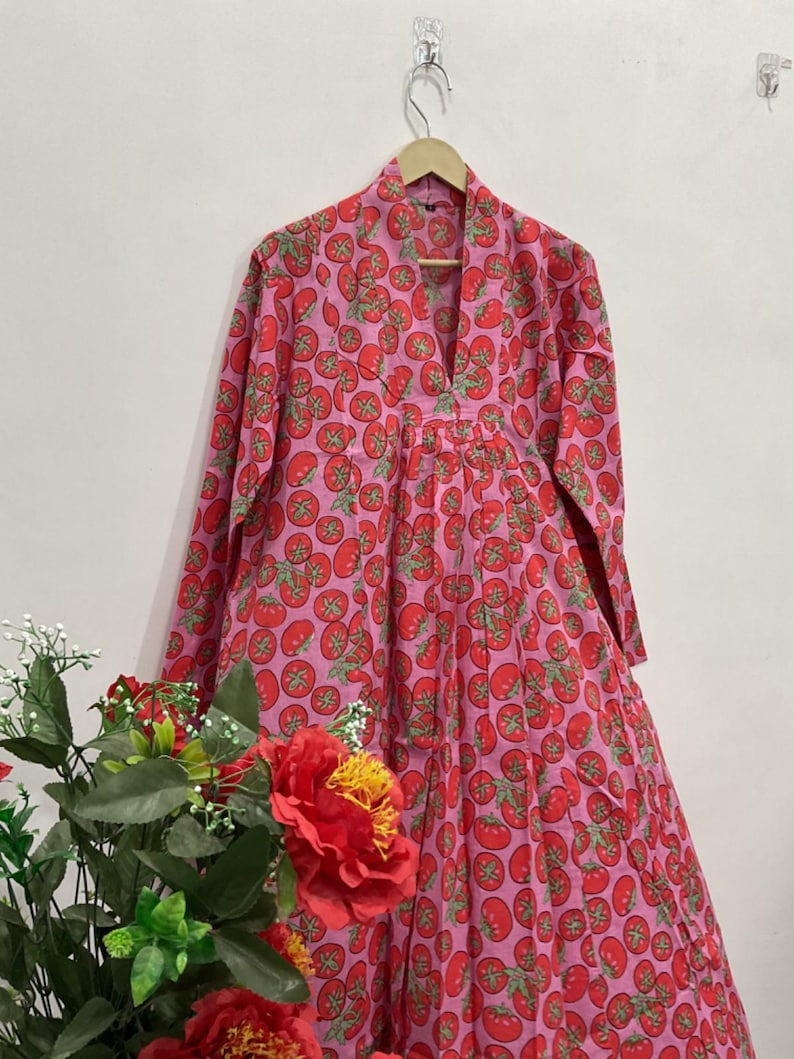 May include: A long-sleeved dress featuring a vibrant pink background with a repeating pattern of red tomatoes and green stems. The garment has a V-neckline, a structured collar, and a pleated bodice that flows into a full-length skirt silhouette.