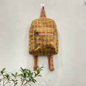 以下が含まれることがあります： 赤と白のアクセントが付いた、黄色の花柄バックパック。 バックパックにはフロントポケットと調節可能なストラップが付いています。 バッグは白い壁に掛けられています。 花柄には赤い花と緑の葉が含まれています。