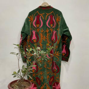 以下が含まれることがあります： ピンク、赤、オレンジの色合いの、精巧な花の刺繍が施されたグリーンのベルベットコート。長いコートは木製のハンガーに掛けられています。コートのデザインは、様式化された花と葉の細かいパターンが特徴です。