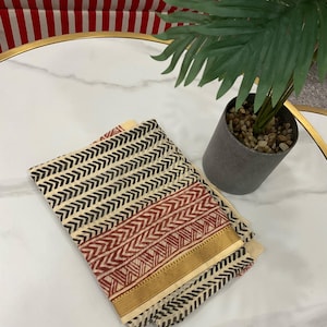 May include: Folded cream-colored fabric with a repeating black and red chevron pattern. A gold border trims the edge. The fabric is on a white marble surface with a small potted plant in the background.