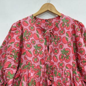 May include: A pink blouse featuring a repeating pattern of red tomatoes with green stems and leaves. The garment has a round neckline with a central tie closure, long sleeves with gathered shoulders, and a loose, flowing fit throughout the bodice.