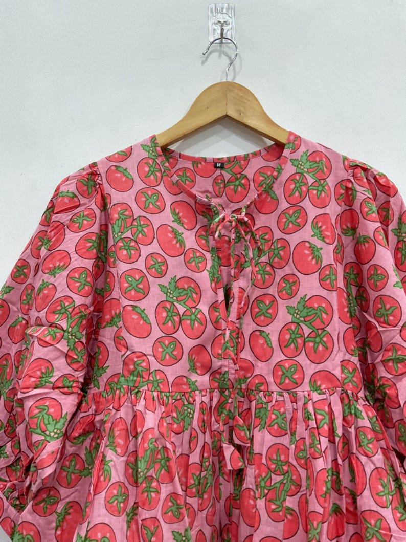 May include: A pink blouse featuring a repeating pattern of red tomatoes with green stems and leaves. The garment has a round neckline with a central tie closure, long sleeves with gathered shoulders, and a loose, flowing fit throughout the bodice.