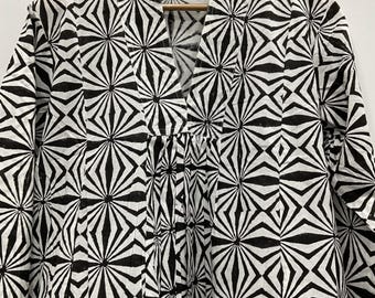 May include: A black and white patterned top with a V-neck design. The geometric pattern features a repeating starburst and diamond design. The top is hanging on a wooden hanger.