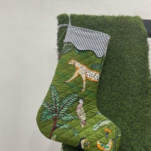 Op de afbeelding: Groene gewatteerde kerstsok met een jungle-thema. De sok heeft een luipaard, papegaaien en tropische planten. De bovenkant is afgewerkt met een zwart-wit gestreepte stof.