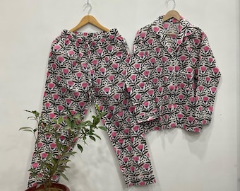 May include: A floral pajama set with a white base and a pattern of pink flowers and green leaves. The set includes a button-down shirt and trousers, both displayed on wooden hangers.