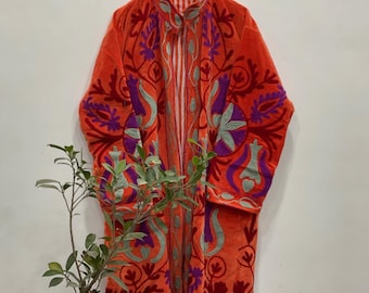 Könnte beinhalten: Ein orangefarbener Samtrobe mit aufwendiger floraler Stickerei in Lila und Türkis. Die Robe hängt an einem Holzbügel vor einer weißen Wand. Eine kleine Topfpflanze steht vor der Robe.