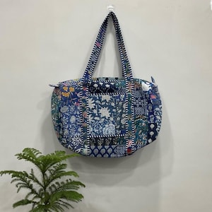 May include: A patchwork tote bag with a blue floral pattern. The bag has two handles and a zipper closure. The bag is hanging on a wall. A small green plant is in the foreground.