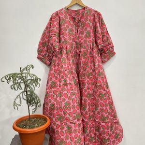 May include: A midi-length dress featuring a vibrant pink background with a repeating pattern of red tomatoes and green leaves. The garment has three-quarter length puff sleeves, a round neckline with a tie closure, and a loose, flowing silhouette.
