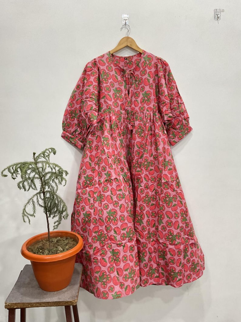 May include: A midi-length dress featuring a vibrant pink background with a repeating pattern of red tomatoes and green leaves. The garment has three-quarter length puff sleeves, a round neckline with a tie closure, and a loose, flowing silhouette.