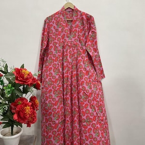 May include: A long-sleeved, floor-length dress in a vibrant pink hue, featuring an all-over repeating pattern of red tomatoes with green stems. The garment has a V-neckline, a button-down front placket, and a loose, flowing silhouette.