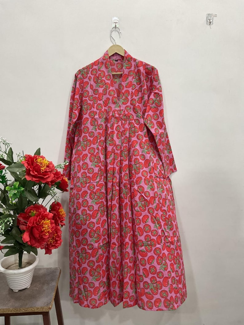 May include: A long-sleeved, floor-length dress in a vibrant pink hue, featuring an all-over repeating pattern of red tomatoes with green stems. The garment has a V-neckline, a button-down front placket, and a loose, flowing silhouette.