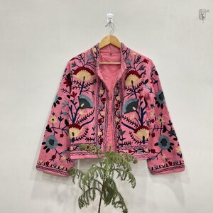 May include: A pink jacket with floral embroidery in shades of blue, yellow, and red. The jacket is hanging on a wooden hanger against a white wall. The embroidery is detailed and covers the front and sleeves of the jacket.