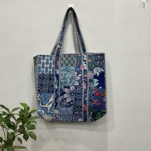 Puede incluir: Un bolso tote patchwork con una variedad de patrones azules y florales. El bolso tiene dos asas y está colgado en una pared blanca. El diseño del bolso incluye varios tonos de azul, blanco y toques de rojo y verde.