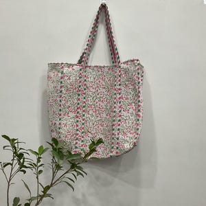 以下が含まれることがあります： ピンクと緑の花柄の白いトートバッグが白い壁に掛けられています。バッグのハンドルはピンクと緑のストライプです。バッグはキルティング生地で作られています。