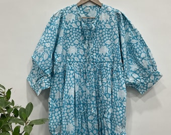 May include: A light blue dress with a white floral pattern. The dress has short, puffy sleeves and a gathered waist. It is hanging on a wooden hanger against a white wall. The dress is made of a lightweight fabric.