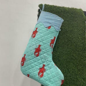 May include: A quilted Christmas stocking with a light blue background and a pattern of red lobsters. The top of the stocking has a blue and white striped fabric with a scalloped edge. A loop is attached for hanging.