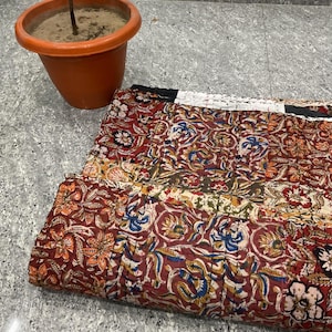 May include: A folded patchwork quilt with a floral pattern in shades of red, orange, blue, and cream. The quilt is made of multiple fabric squares stitched together. A terracotta pot with a small plant is in the background.