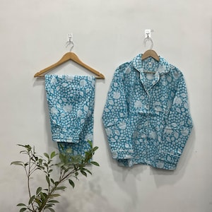 Puede incluir: Un conjunto de pijama de dos piezas en un estampado floral azul claro y blanco. El conjunto incluye una camisa con botones y pantalones a juego, ambos colgados de perchas de madera contra una pared blanca. Los pijamas tienen un ajuste relajado.
