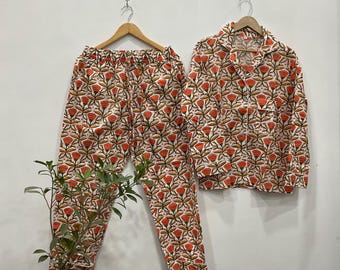 May include: A matching pajama set with an orange and brown floral design on a white backdrop. The set comprises a collared, button-down shirt and trousers. The pajamas are displayed on wooden hangers.